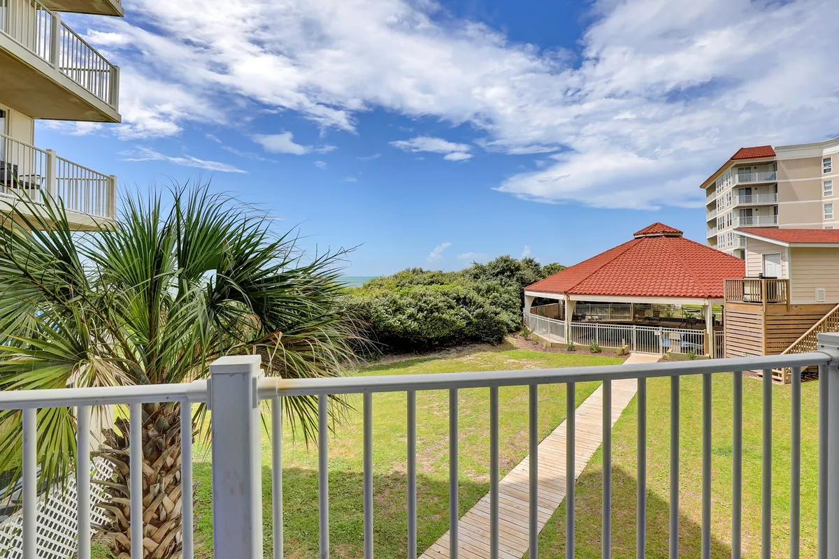 The image showcases a tranquil beachfront scene at North Topsail Beach, with a wooden walkway leading guests through swaying palm trees to a charming red-roofed gazebo overlooking the ocean. The soft sand, serene blue water, and inviting pathway evoke relaxation and a sense of coastal escape.