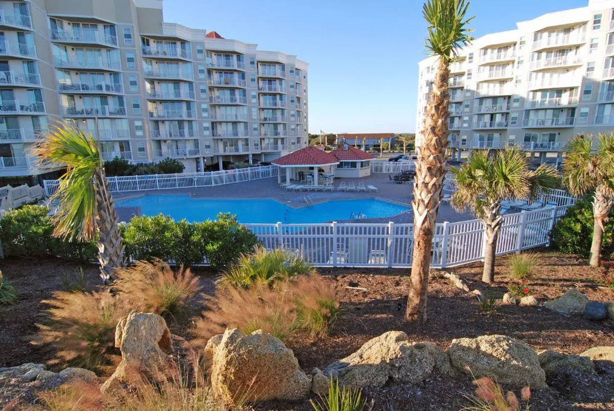 A spacious swimming pool surrounded by palm trees and residential resort buildings, with a landscaped garden creating a lush, inviting atmosphere. The setting offers ample space for swimming, sunbathing, and group gatherings by the pool.