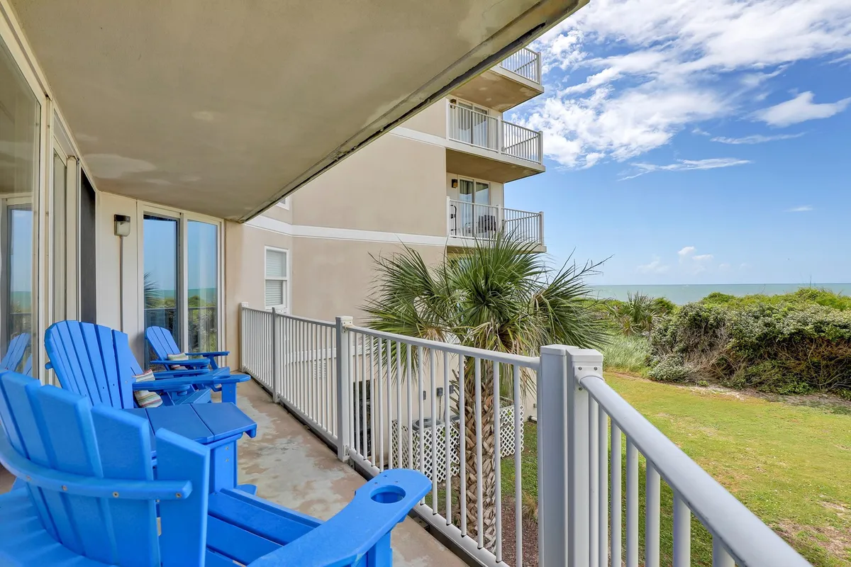 This exterior image features blue Adirondack chairs arranged on a balcony, overlooking a breathtaking ocean view framed by palm trees and a balcony railing. The scene is tranquil, suggesting a private space perfect for morning coffee, evening relaxation, or gazing at the sunrise and sunset.