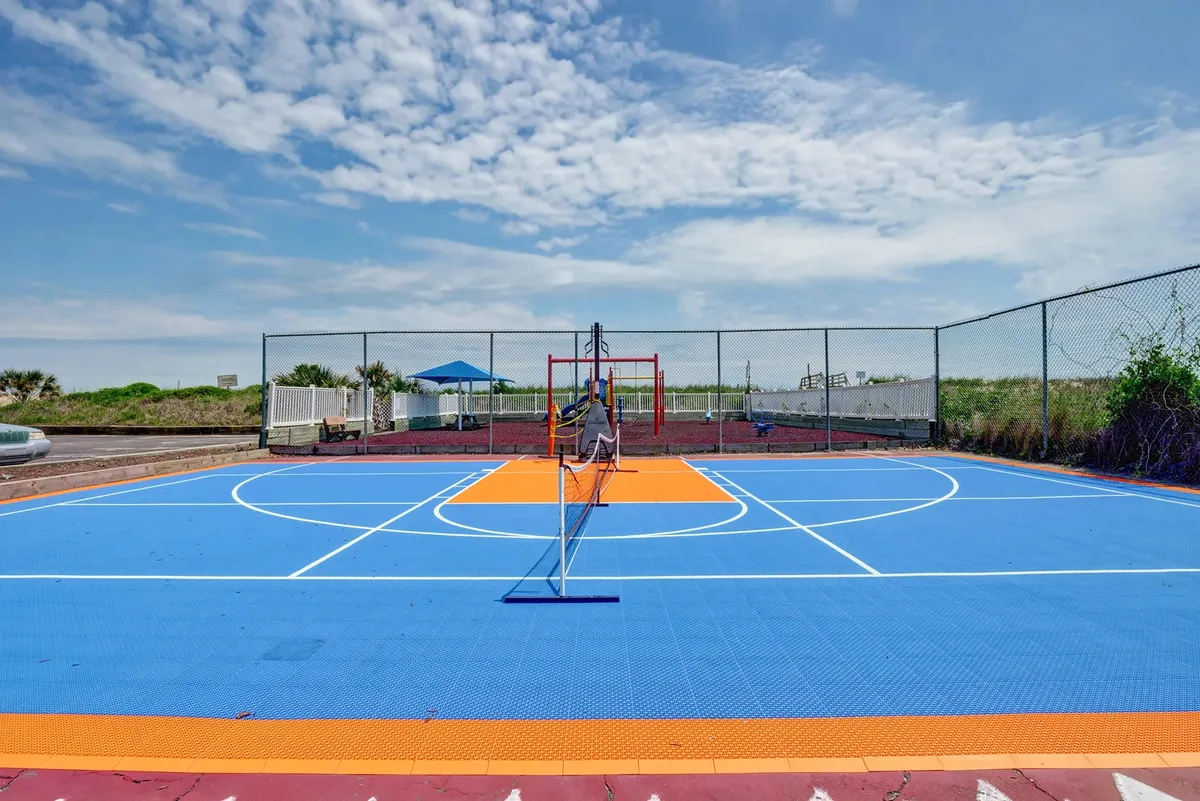 An energetic outdoor sports area with a colorful basketball court, tennis net, adjacent play area, and ample seating surrounded by greenery. Guests can be seen enjoying games and socializing under sunny skies.