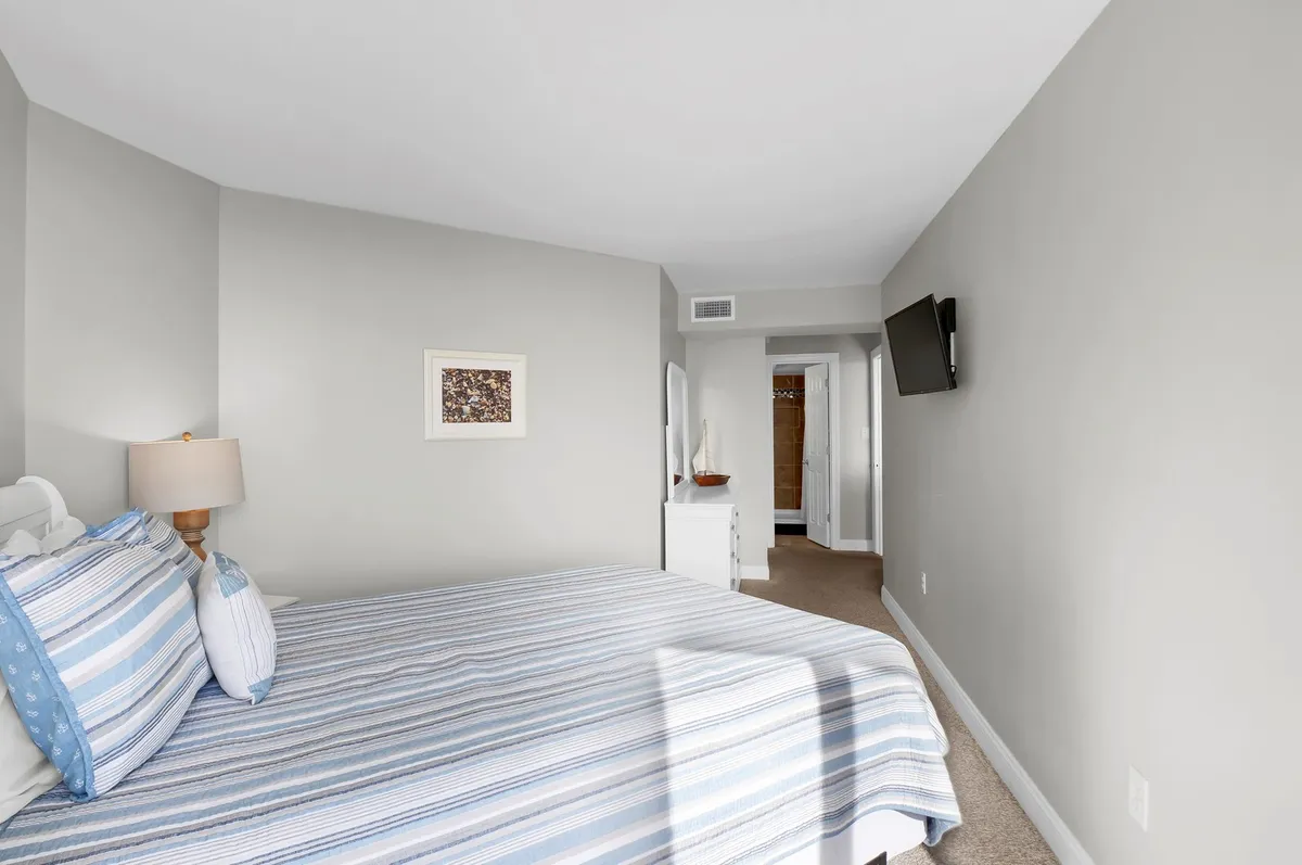 A comfortable bedroom featuring striped bedding, a lamp on the nightstand, framed artwork, and a television mounted on the wall. The room is softly lit, emphasizing a serene and inviting atmosphere.