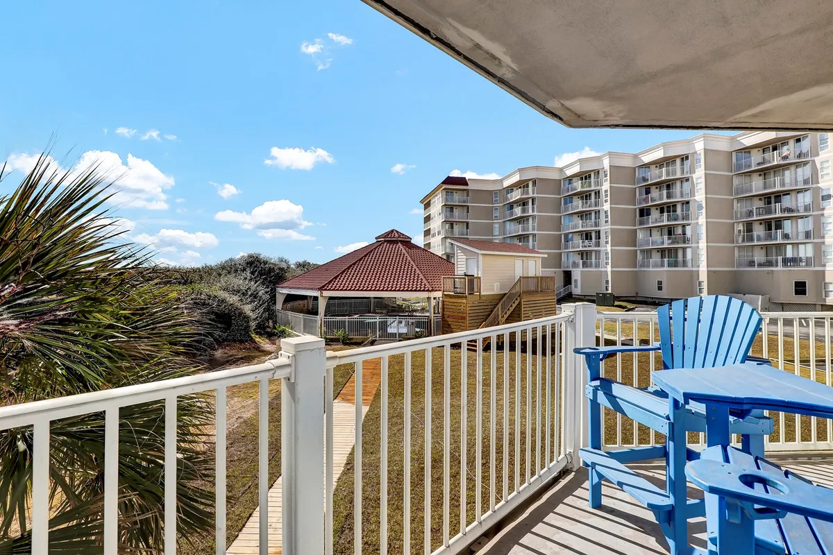 A tranquil exterior scene featuring blue Adirondack chairs on a private balcony, a red-roofed gazebo ideal for gatherings, and modern condominium buildings surrounded by lush greenery. The setting invites groups to relax outdoors, share meals, and enjoy the coastal atmosphere together.