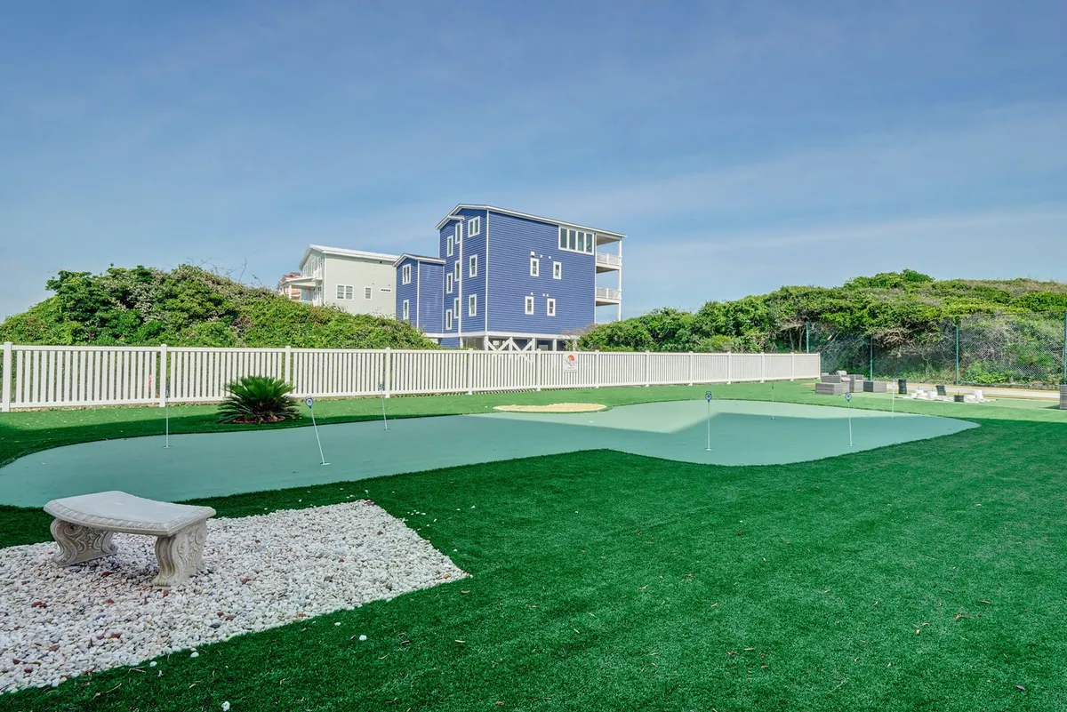 An exterior view of a beautifully maintained artificial putting green surrounded by lush greenery and a stone bench, with a charming blue house in the background. The scene suggests a leisurely atmosphere ideal for golf practice or relaxing outdoors.