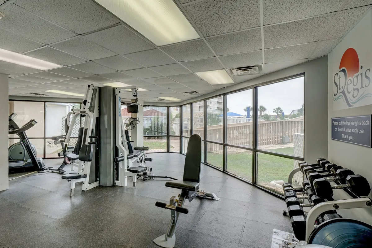 A bright, modern fitness center with large windows letting in natural light, exercise machines, free weights, and visible gym signage. The atmosphere is motivational and energetic, inviting guests to prioritize wellness.