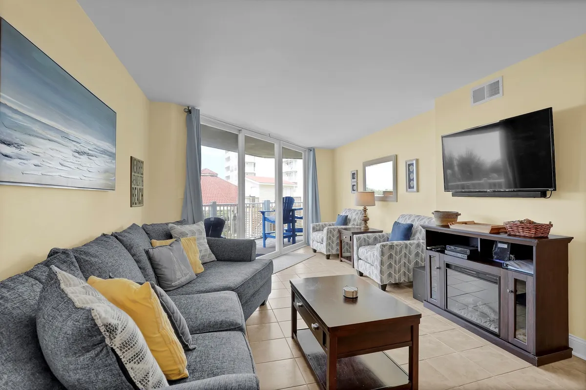 A sun-filled living room featuring a gray sectional sofa, a wooden coffee table, large ocean-facing windows that let in natural light, a TV, and decorative art pieces. The space is set up for relaxing, watching TV, and socializing, and the windows offer glimpses of the sparkling water outside.